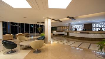 A modern hotel lobby with a reception desk, seating area, and large windows.