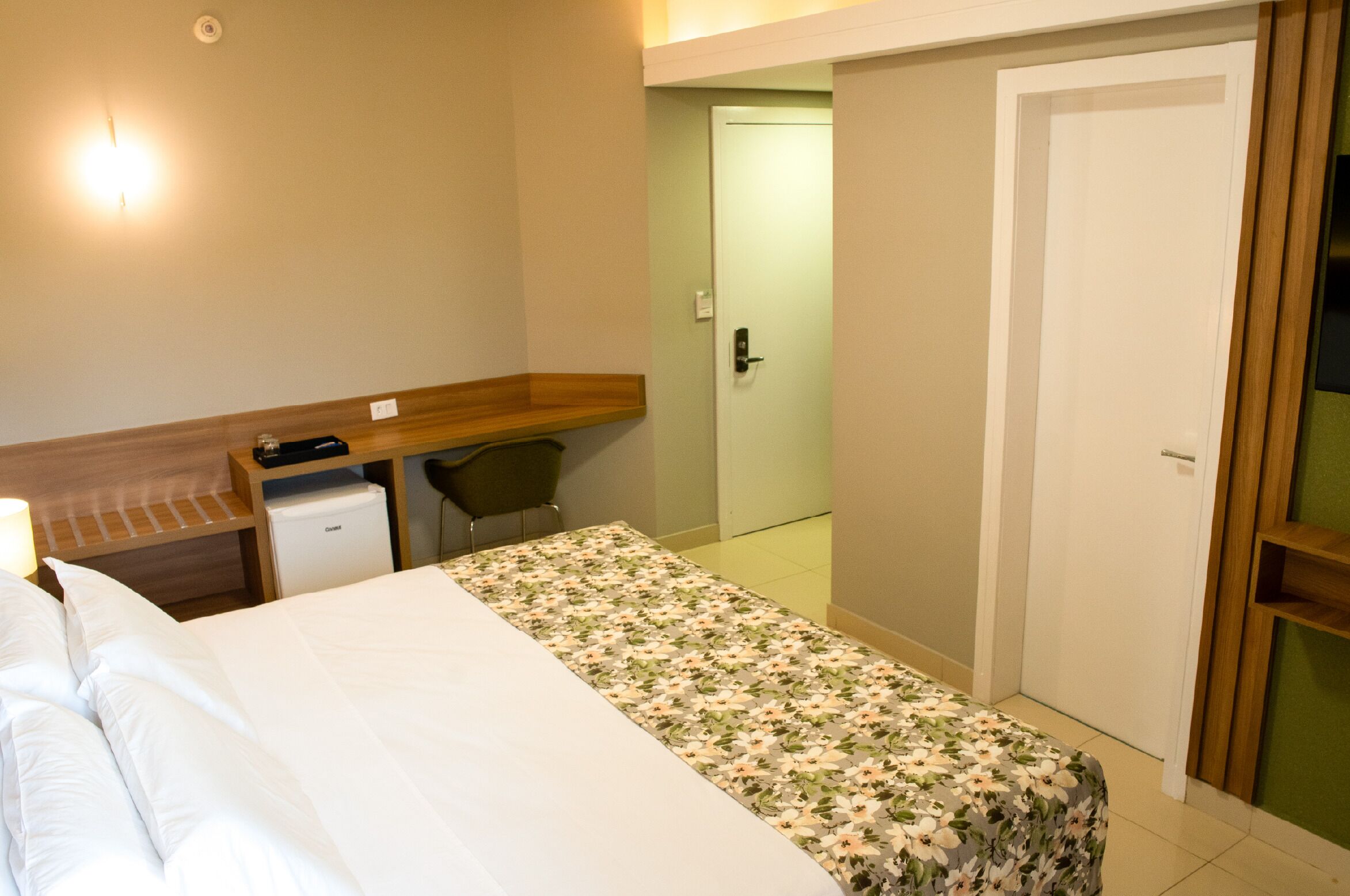 Quarto de hotel com uma cama, uma escrivaninha com cadeira, um pequeno frigorífico e uma porta.
