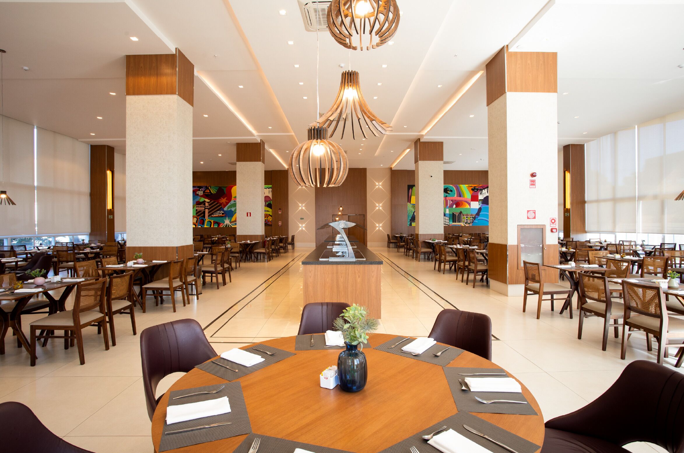 Ampla sala de jantar de hotel com várias mesas, um balcão de recepção central e iluminação pendente moderna.