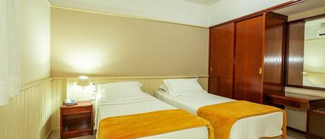 A hotel room with two beds, a wooden wardrobe, a bedside table with a lamp, and a large mirror.