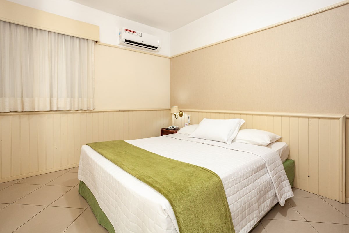 Une chambre d’hôtel comprenant un lit, une table de chevet, un appareil de climatisation, une fenêtre avec des rideaux et une lampe fixée au mur.