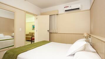 Una habitación de hotel con cama, una manta verde, una mesita de noche con lámpara, un equipo de aire acondicionado y una puerta que da a otra habitación.