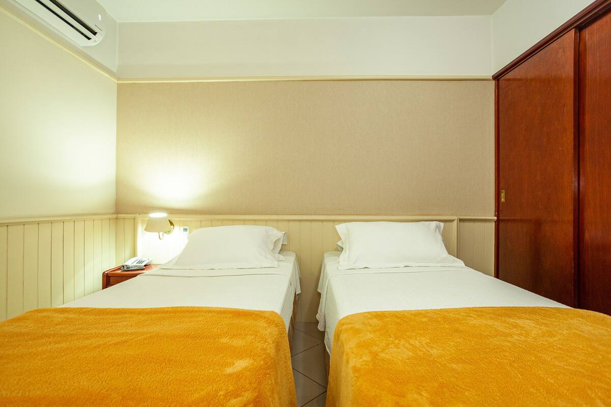 Une chambre d’hôtel avec deux lits simples, une armoire en bois, une table de chevet avec une lampe et un climatiseur fixé au mur.