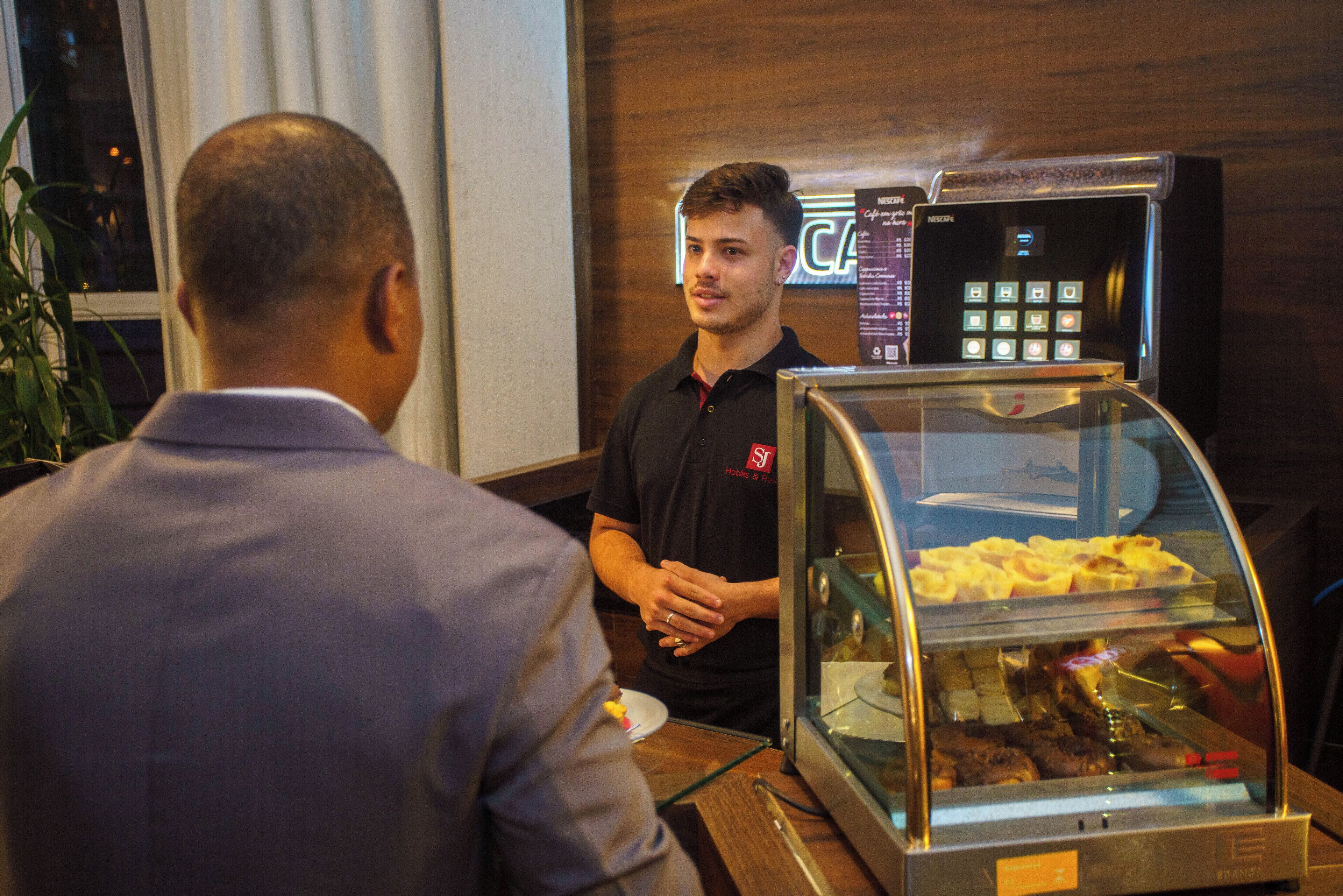 A man in a black shirt standing behind a glass display case with food items, in a room with wooden walls and a window.