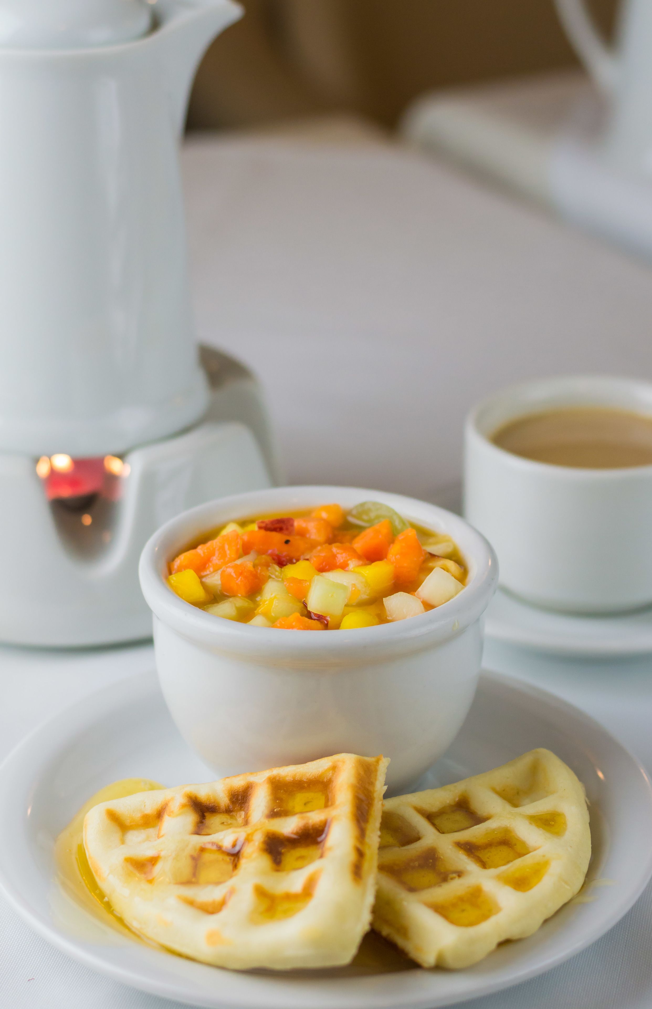 A white teapot, a bowl of fruit soup, a cup of coffee, and two waffles on a white table.
