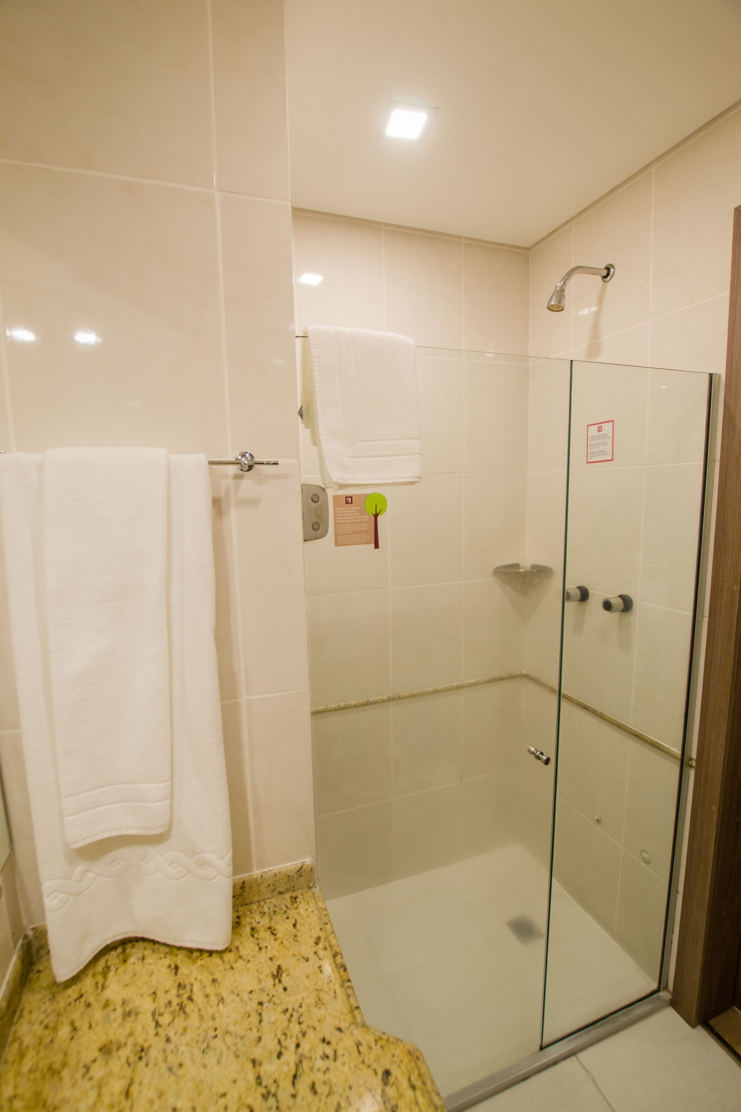 Um chuveiro com paredes de vidro, uma toalha branca pendurada em um porta-toalhas, uma porta de vidro e piso revestido com azulejos.