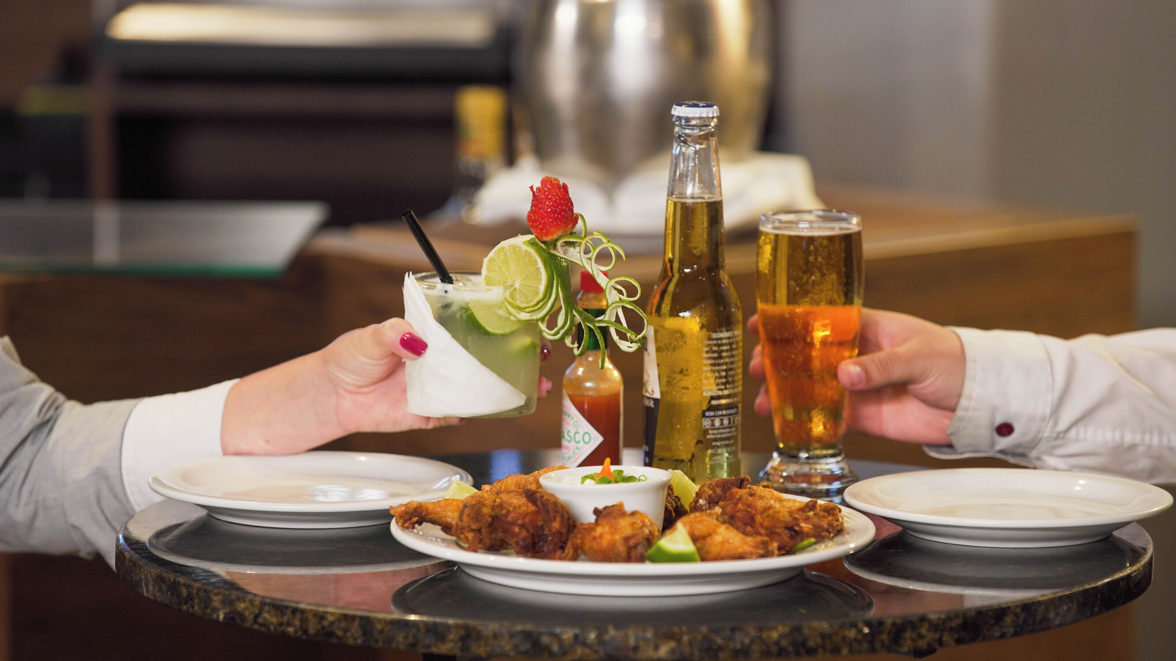 A dining table with plates of fried chicken, a cocktail with a lime garnish, and two bottles of beer.