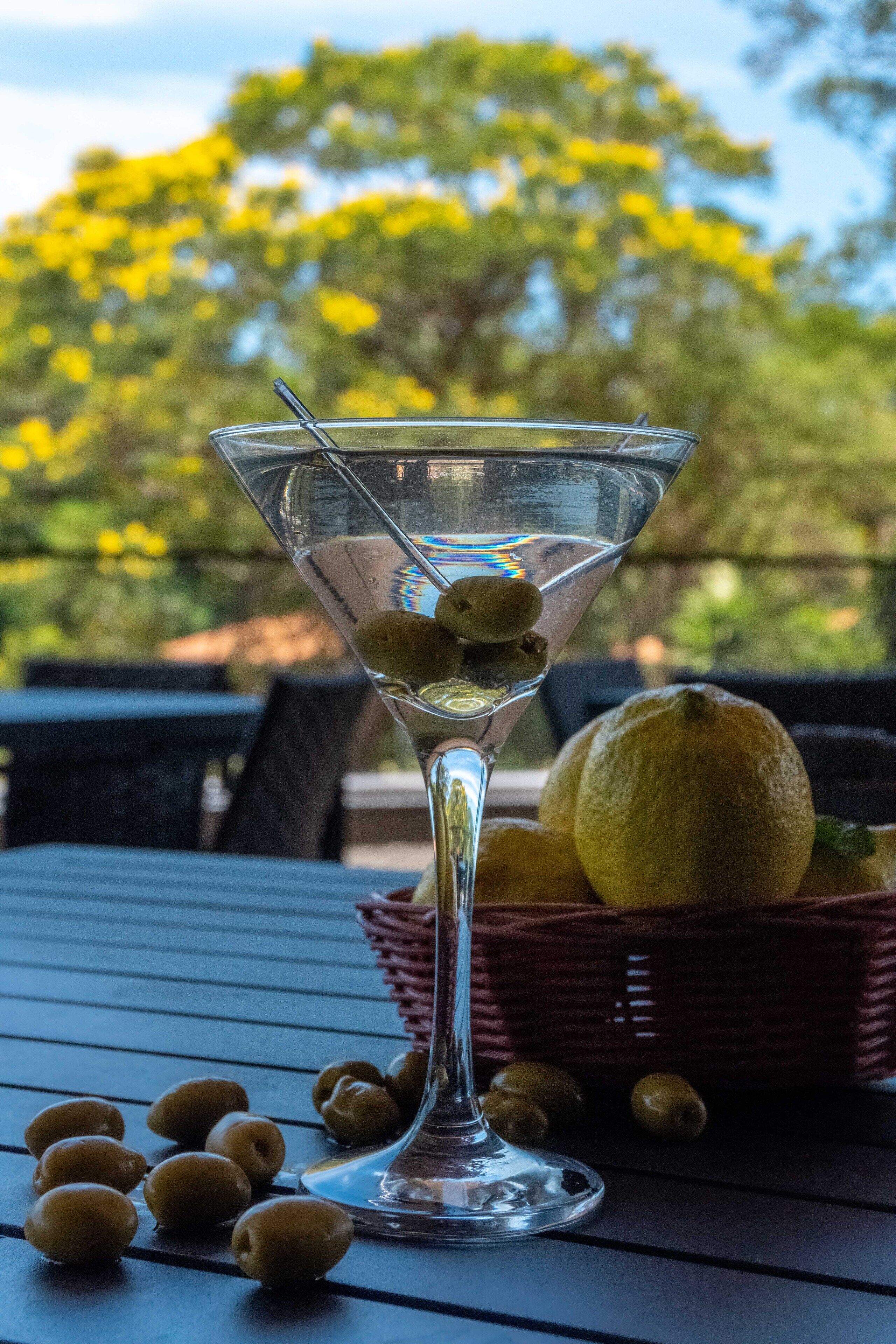 A martini glass with olives on a table with a wicker basket and lemons in the background.