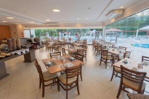 A spacious indoor dining area with multiple tables and chairs, large windows offering a view of an outdoor pool area. -  Complexo Eco Cataratas Resort by SJ (Foz do Iguaçu)