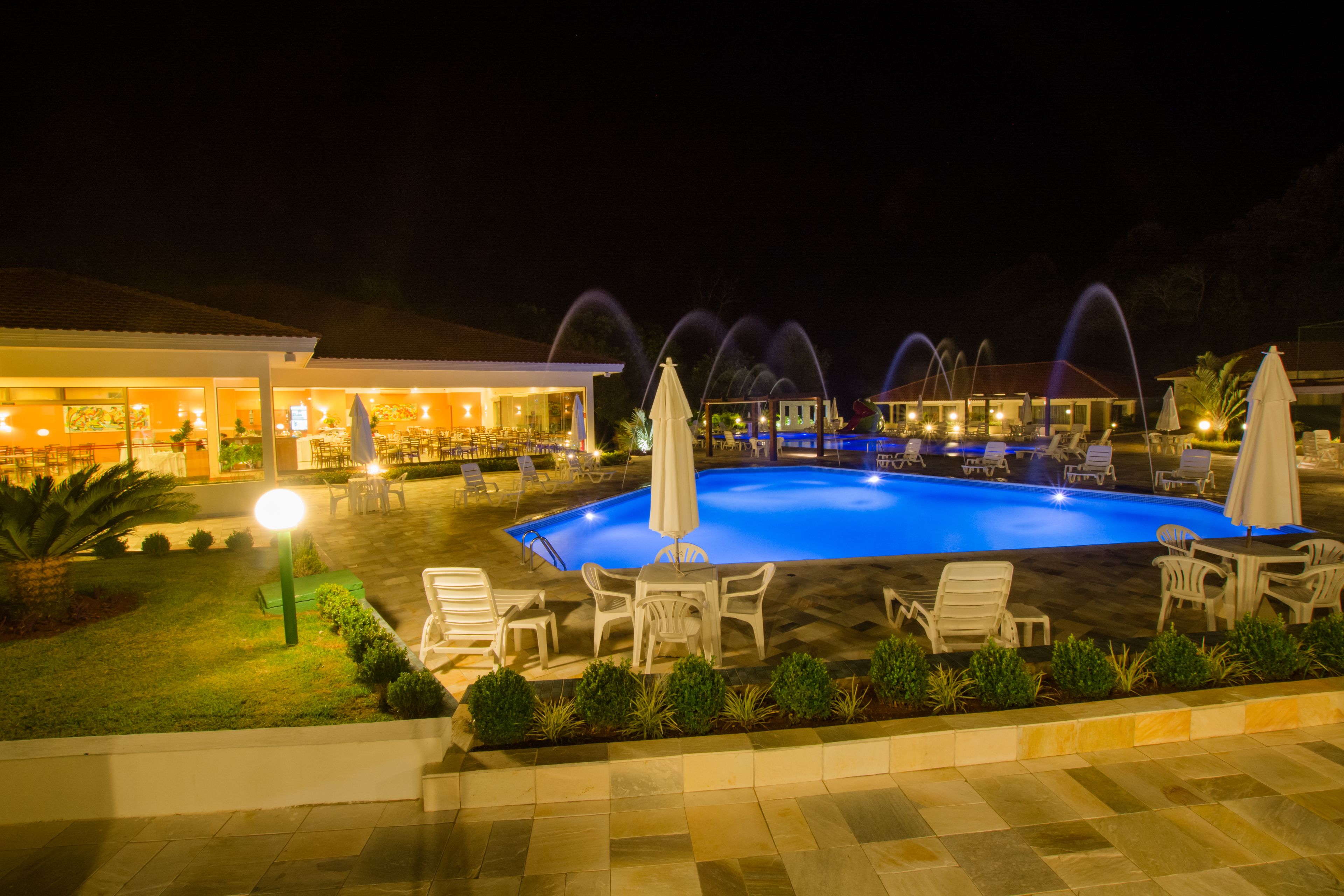 A nighttime swimming pool area with lounge chairs and umbrellas, surrounded by a well-lit building with visible windows and a patio.