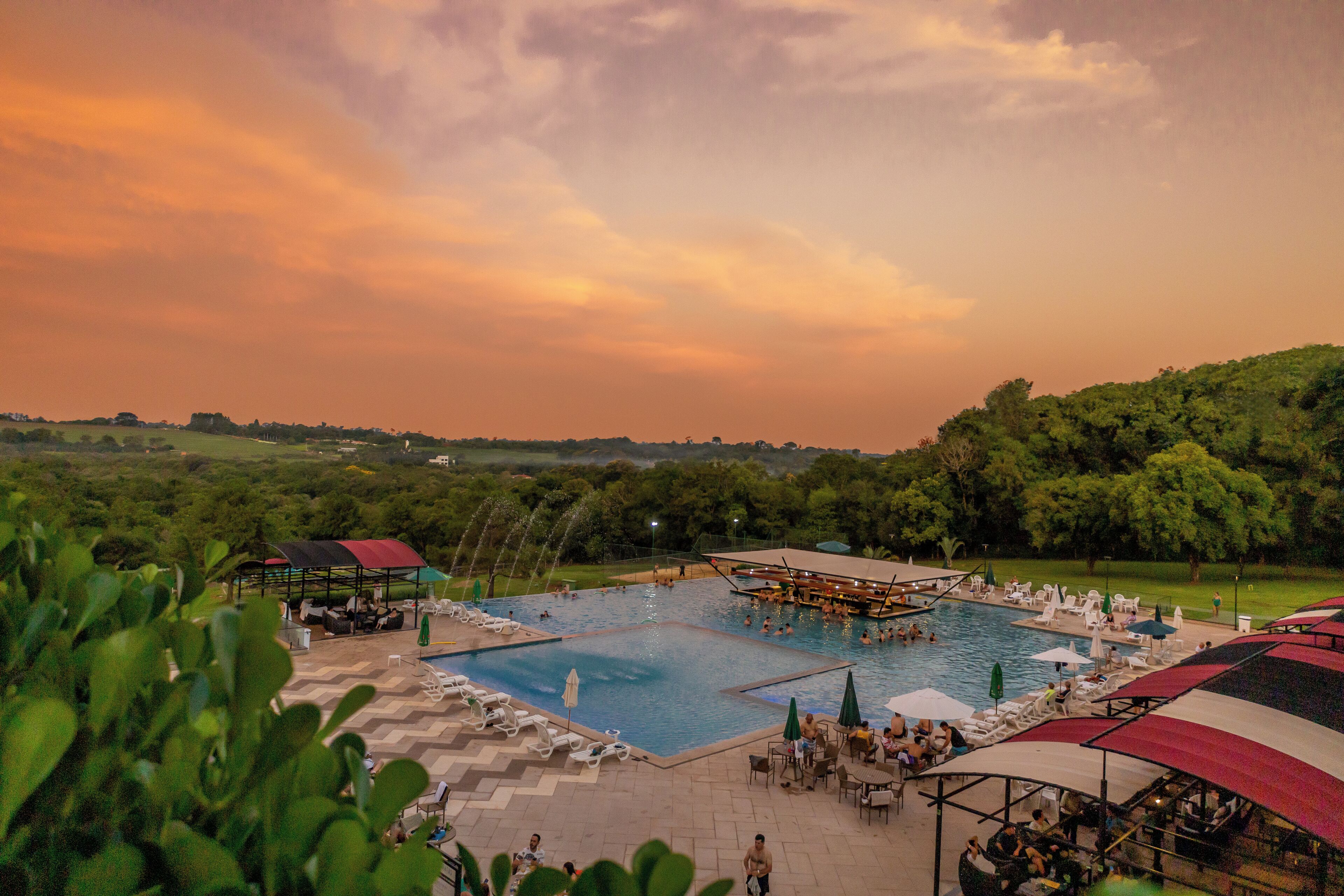 Une grande piscine extérieure divisée en plusieurs zones, entourée de chaises longues et de parasols, avec en arrière-plan une végétation luxuriante et un ciel au coucher du soleil.
