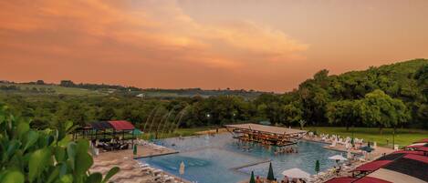 Une grande piscine extérieure divisée en plusieurs zones, entourée de chaises longues et de parasols, avec en arrière-plan une végétation luxuriante et un ciel au coucher du soleil.