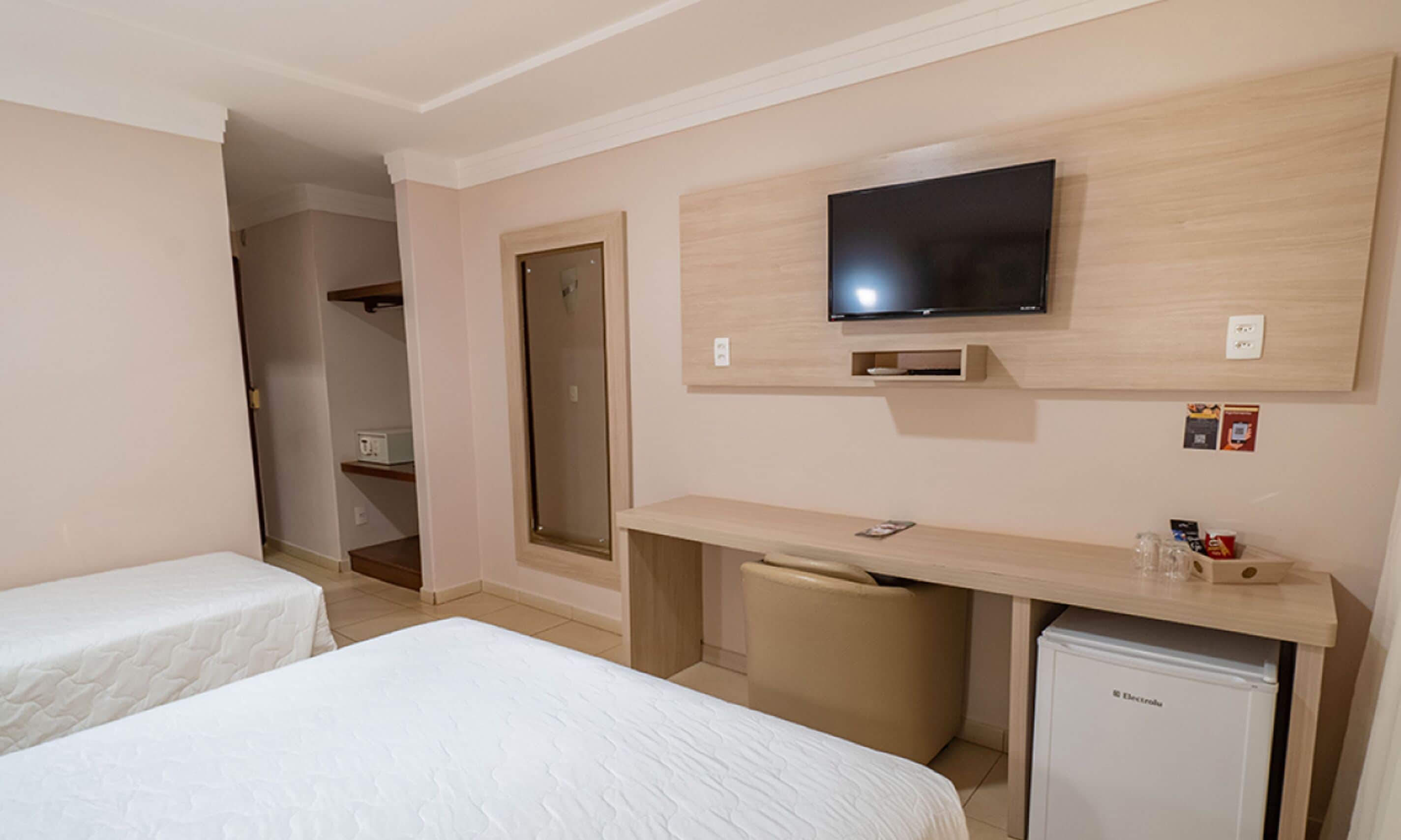Hotellihuone, jossa on kaksi sänkyä, seinälle asennettu televisio, pieni jääkaappi ja työpöytä tuolilla.