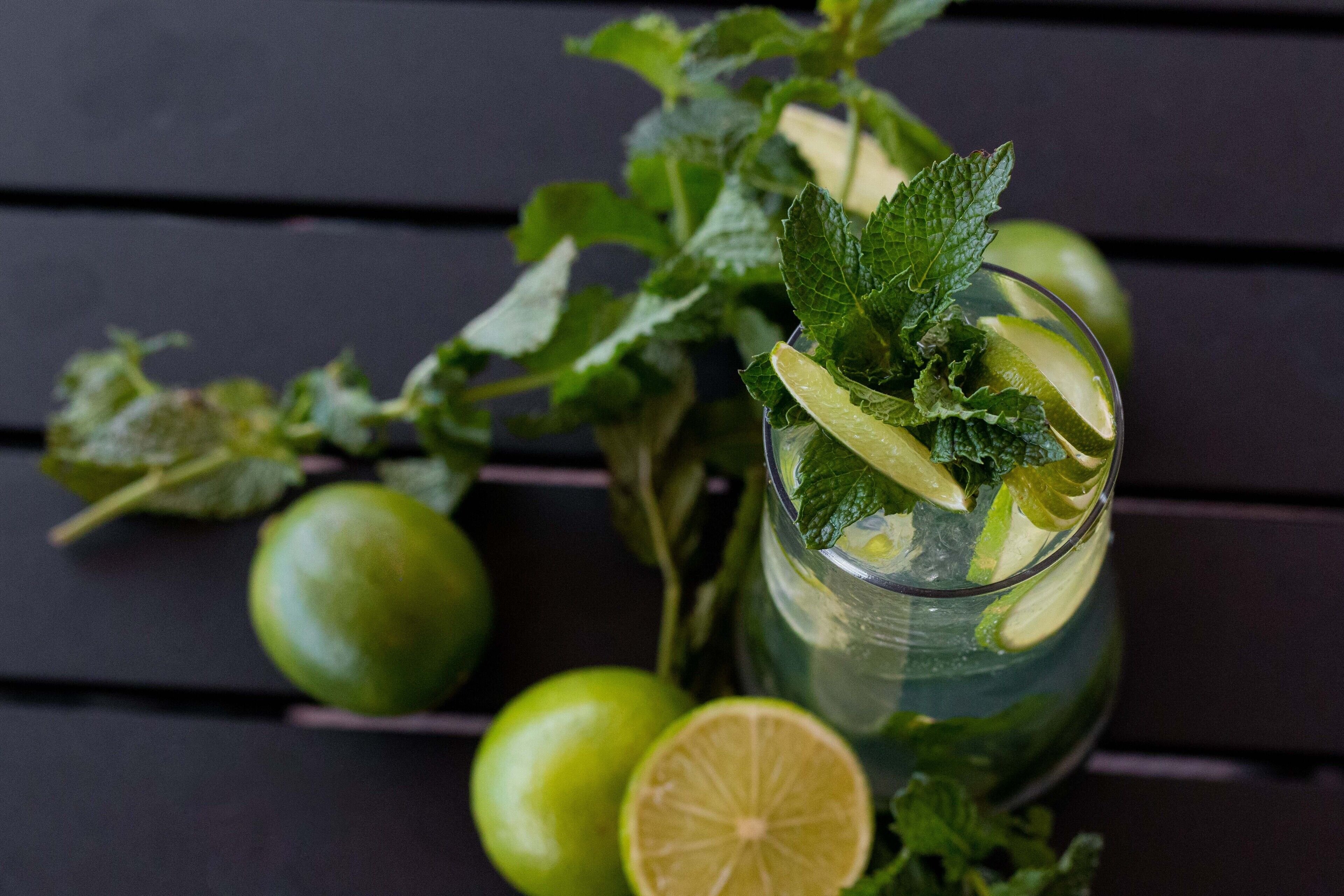 Un vaso de bebida con lima y menta, rodeado de limas enteras y en rodajas sobre una superficie de madera oscura.