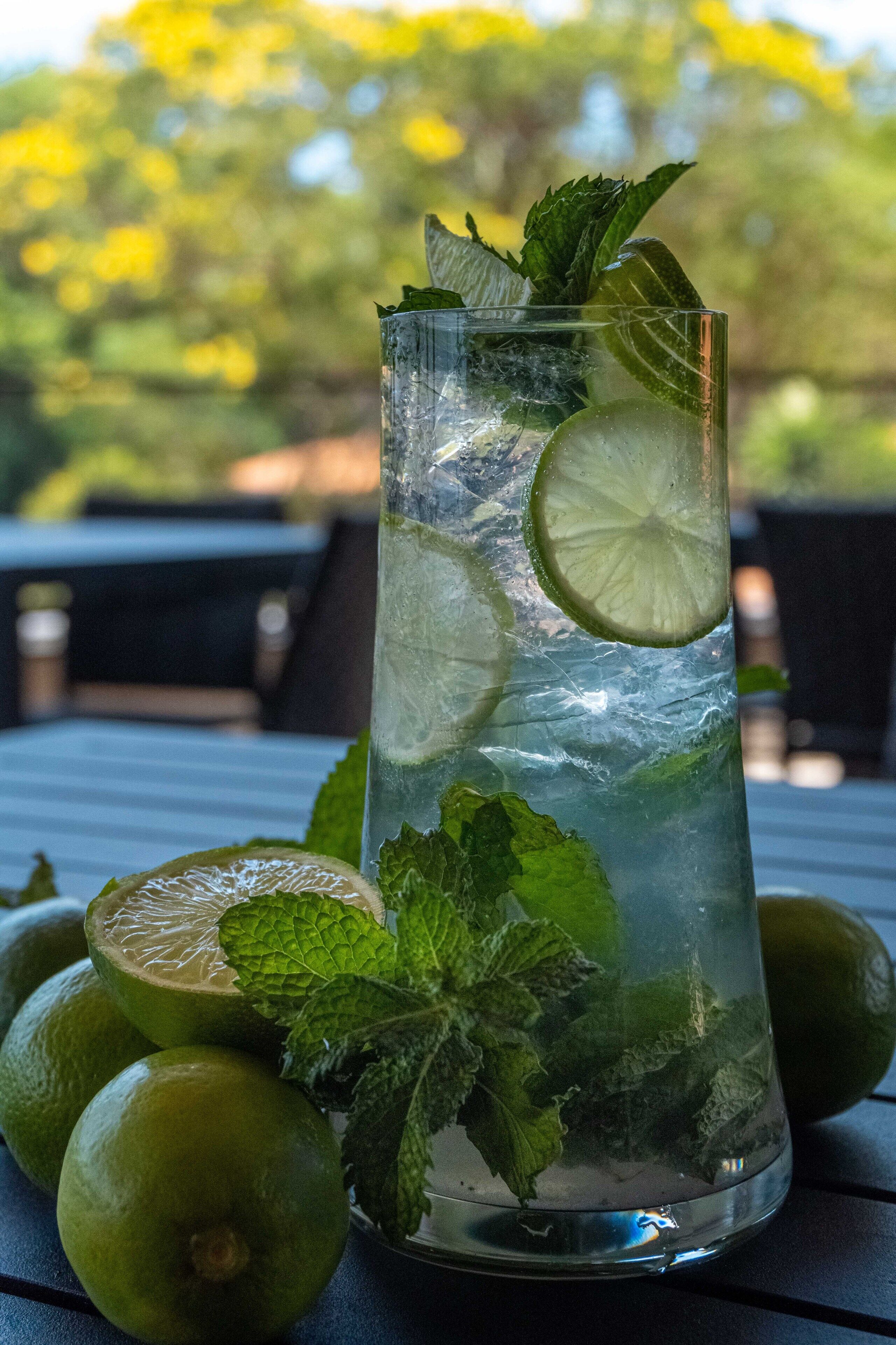 A tall glass filled with a clear liquid, ice cubes, lime slices, and mint leaves, placed on a table with additional limes and mint leaves nearby.