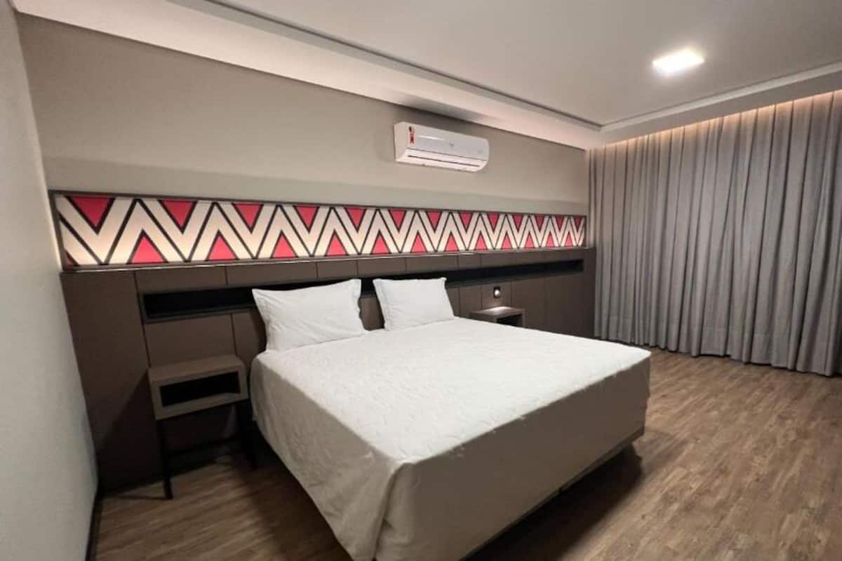 Une chambre d’hôtel moderne avec un grand lit, une table de chevet, une petite table d’appoint, un climatiseur mural et une tête de lit à motifs.