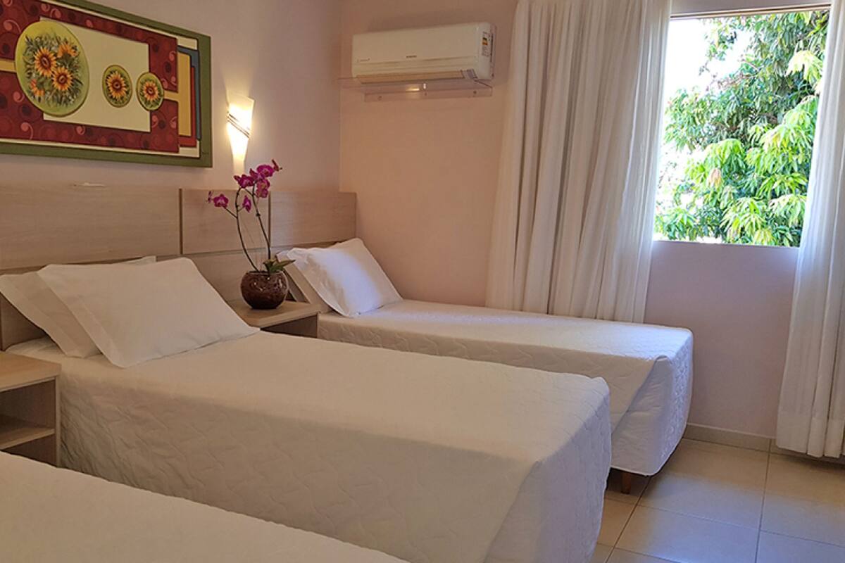 Une chambre d’hôtel avec deux lits simples, une table de chevet avec un téléphone, un élément décoratif mural, un appareil de climatisation et une fenêtre avec des rideaux.
