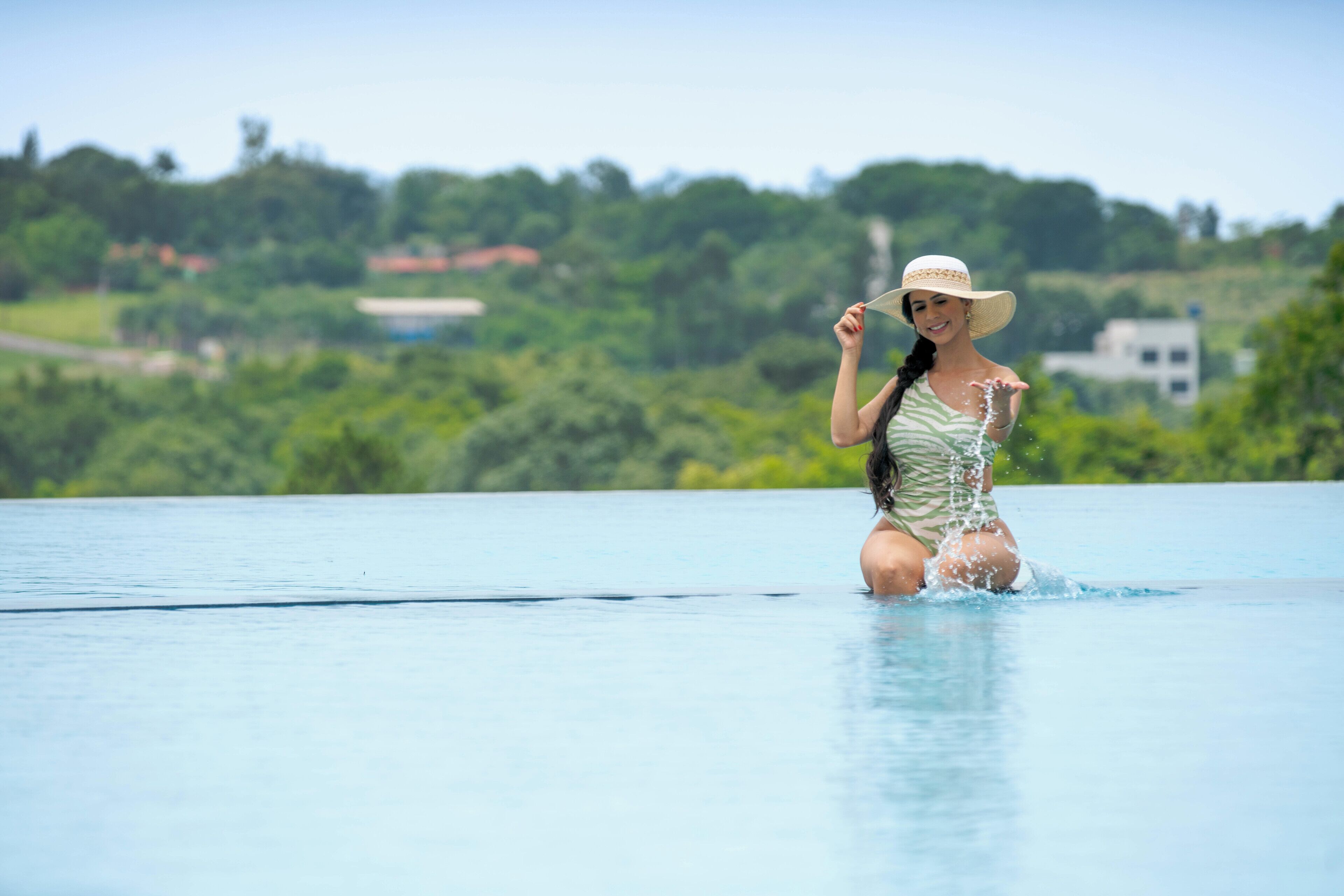 Une femme en maillot de bain et portant un chapeau, debout dans une piscine, avec en arrière-plan une vue sur la verdure et des bâtiments.
