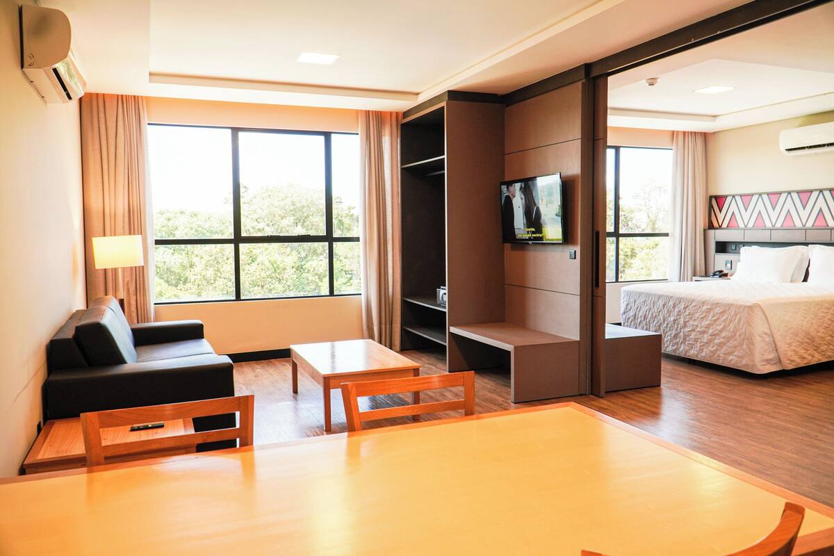 Une chambre d’hôtel avec un canapé, une table, une télévision, un lit et une vue sur la verdure environnante.