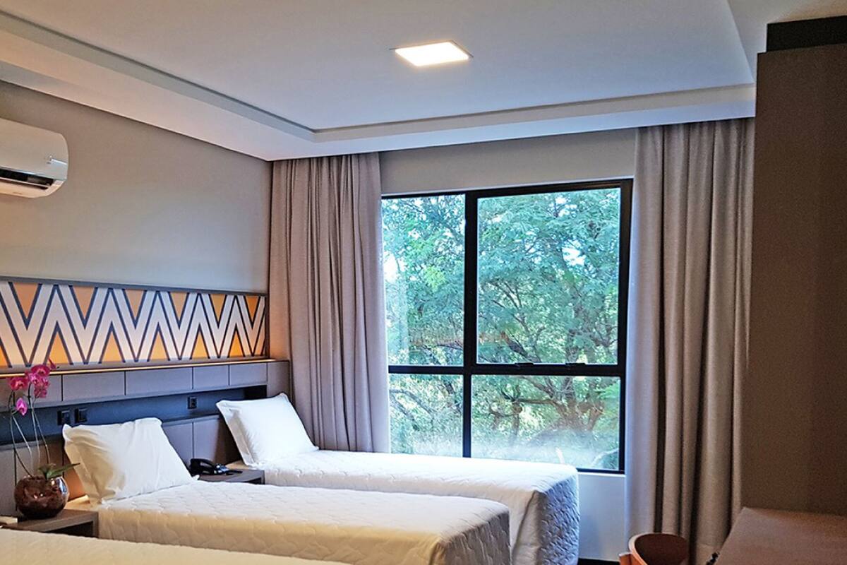 Une chambre d’hôtel avec deux lits, une grande fenêtre donnant sur la verdure, un téléviseur à écran plat et un panneau décoratif mural.