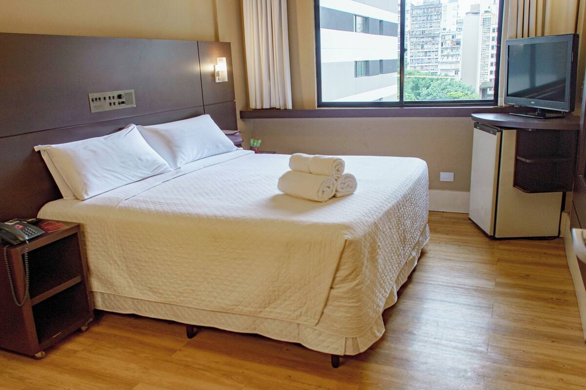 Een net opgemaakt bed met witte lakens, een nachtkastje met een telefoon en een televisie op een standaard.
