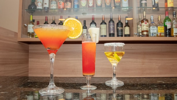 Tres cócteles sobre una barra con una estantería de botellas de licor al fondo.