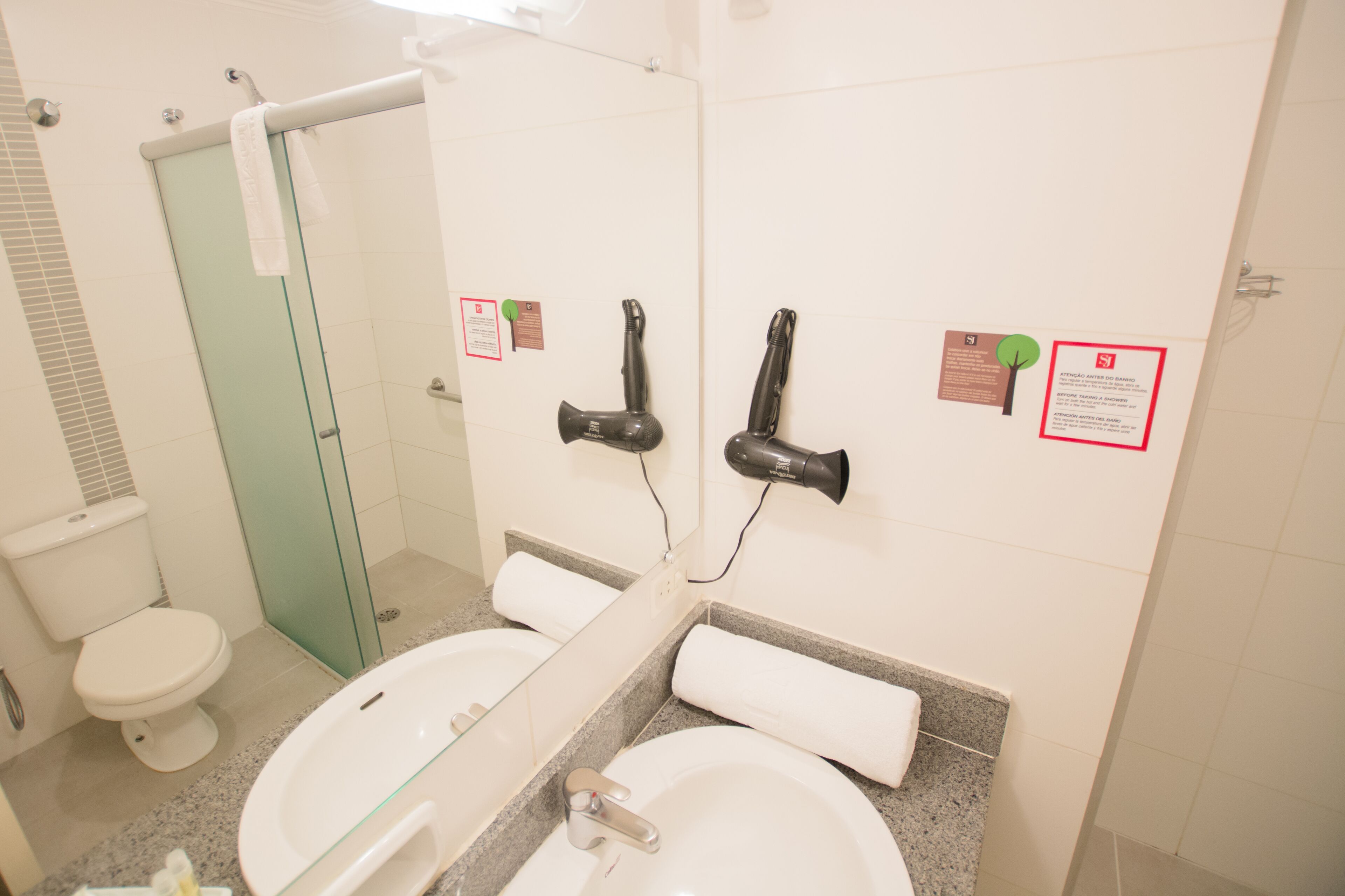 Une salle de bain avec une douche à parois de verre, des toilettes blanches, un lavabo avec un miroir et deux sèche-cheveux fixés au mur.