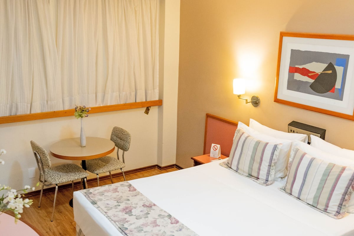 Ett hotellrum med en säng, ett runt bord, två stolar, ett litet bord med en vas och en väggmonterad lampa.