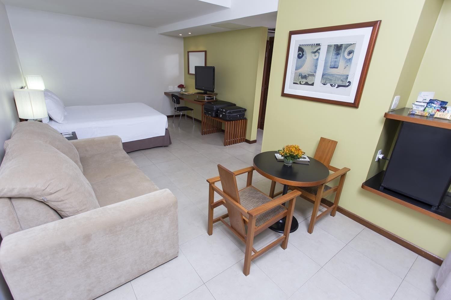 Habitación de hotel con cama, sofá, mesa de comedor con sillas, televisor y un cuadro en la pared.
