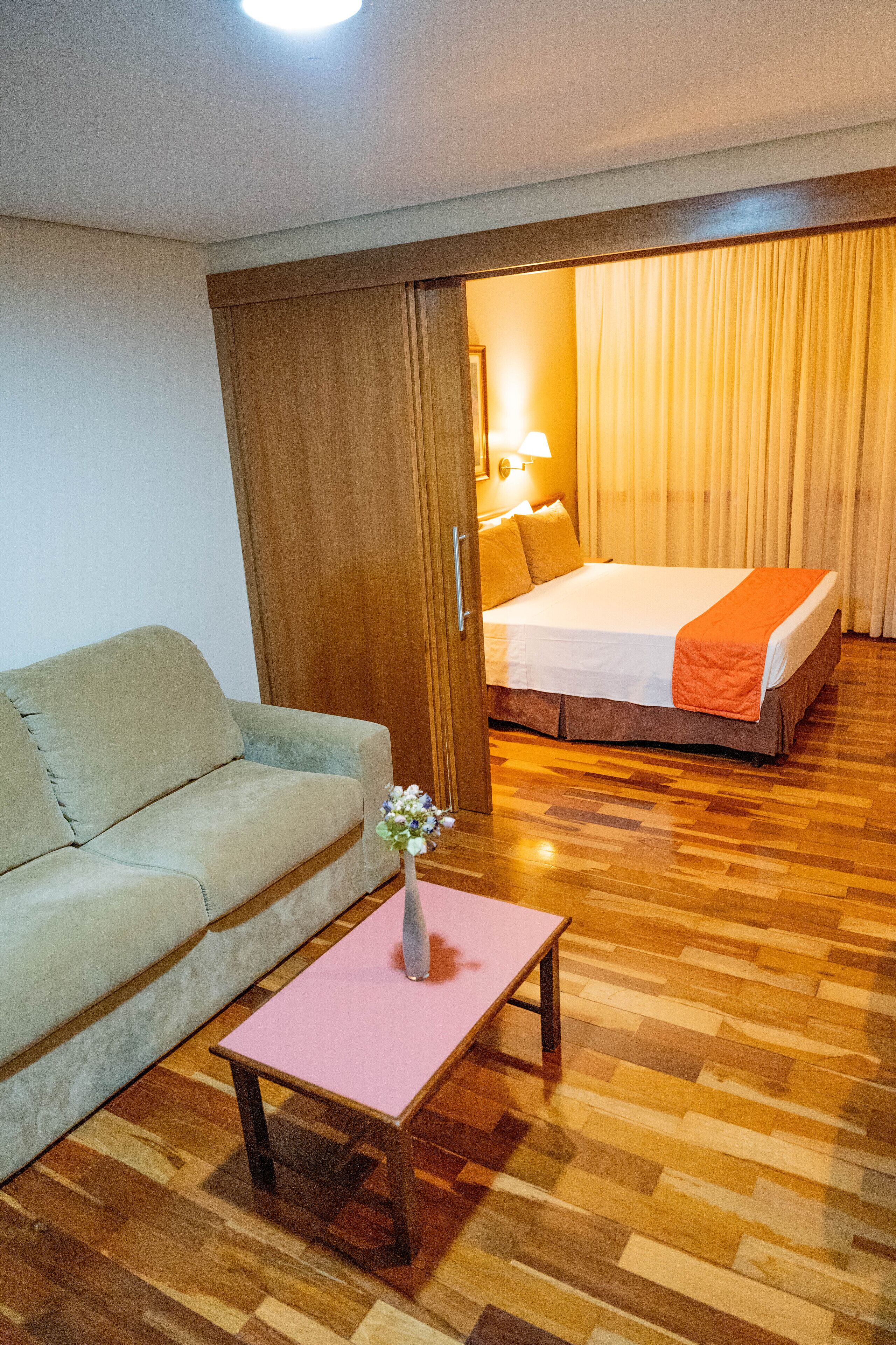 Een hotelkamer met een bed, een slaapbank, een klein tafeltje met een vaas, houten vloeren en een houten deur.