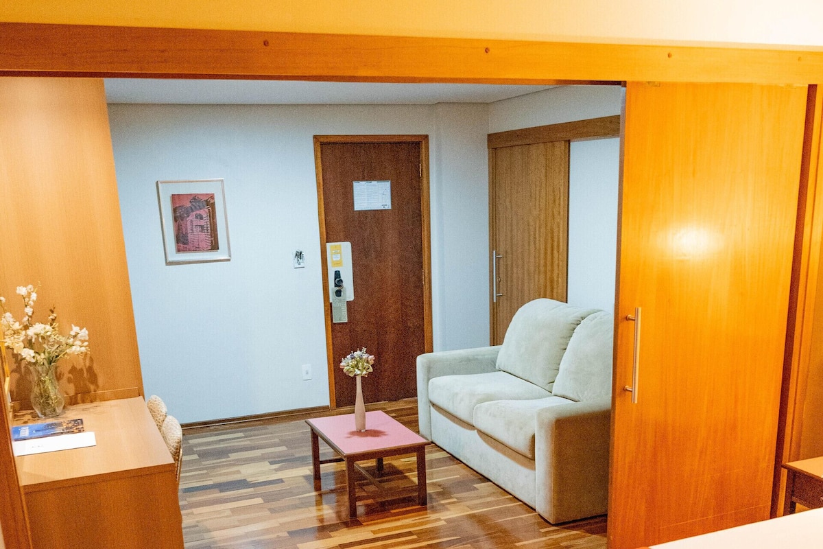 Ett hotellrum med trägolv, en säng, ett skrivbord, en stol, en soffa och ett litet bord med en vas.
