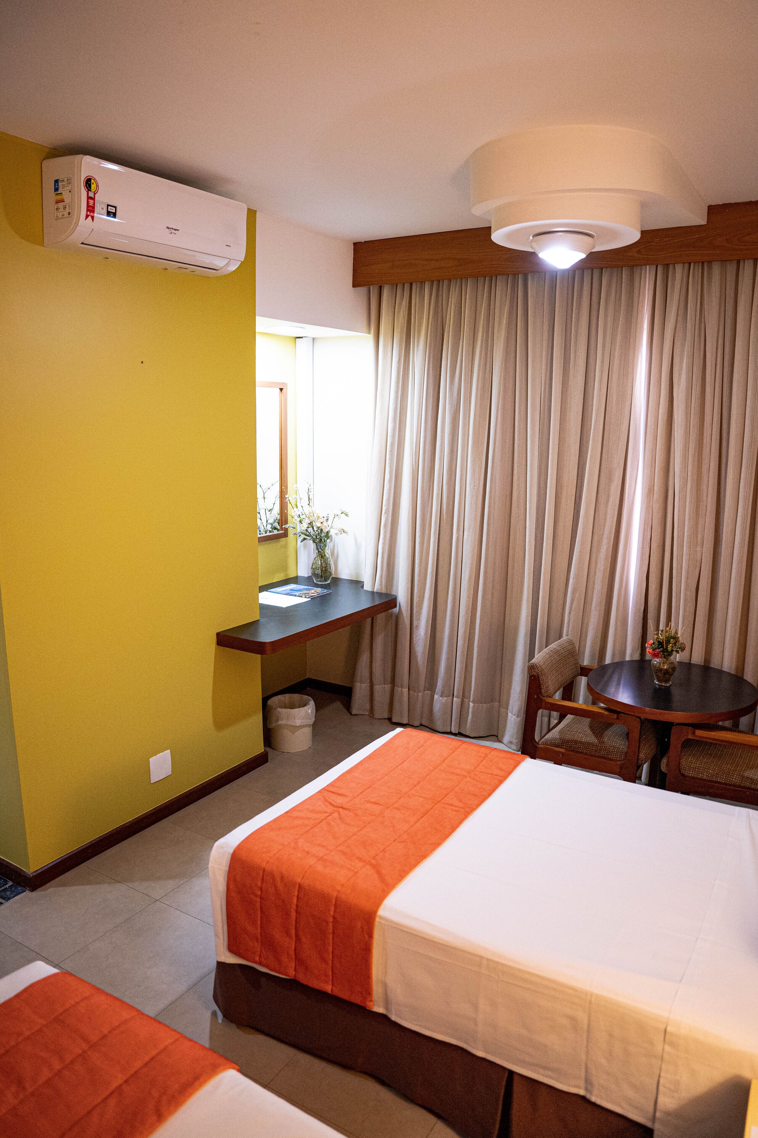 A hotel room with two beds, a desk, a chair, a table with a vase, and a window with curtains.