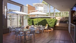 Un patio al aire libre con mesas y sillas de metal, sombrillas y vistas a un edificio con escaleras.