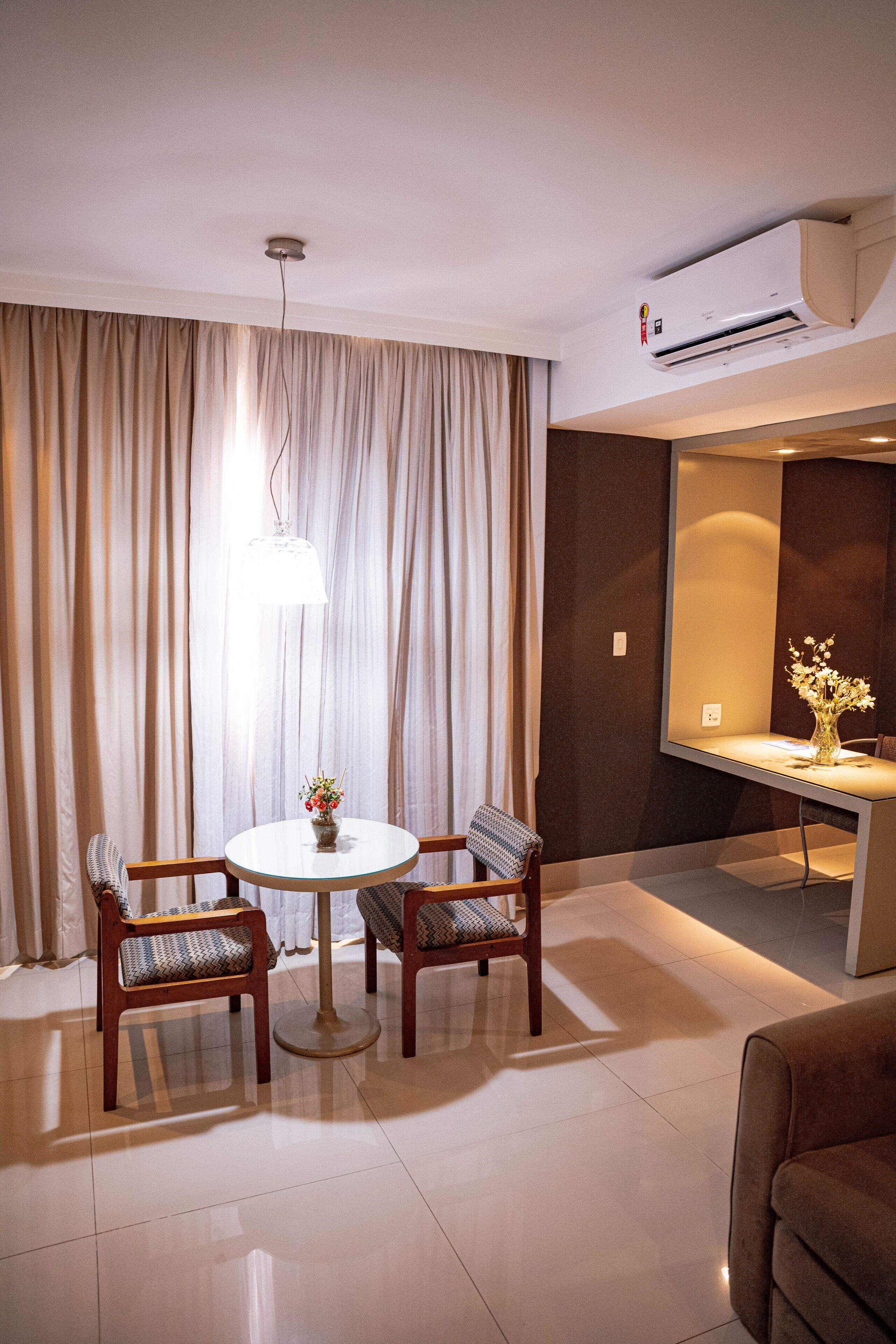 Une chambre d’hôtel moderne avec une table à manger ronde, deux chaises, un canapé, un bureau agrémenté d’un vase de fleurs et une grande fenêtre avec des rideaux.