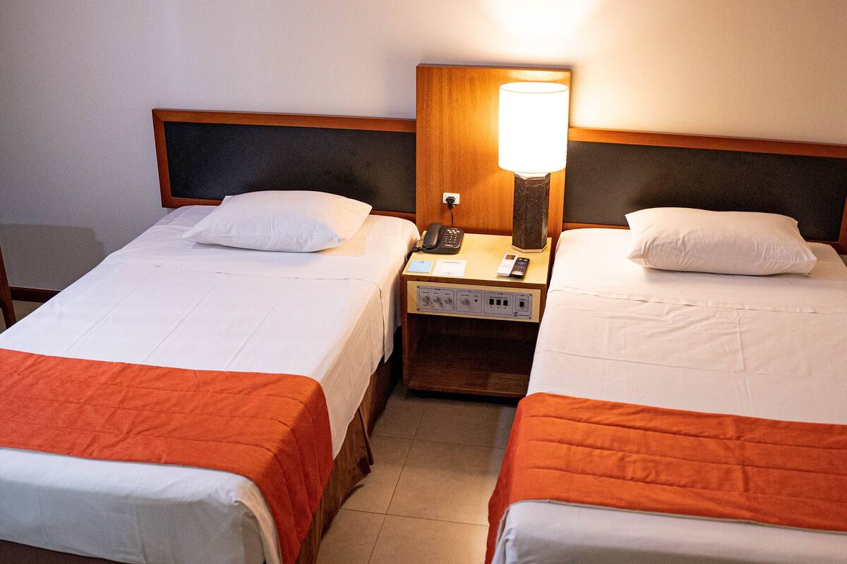 Ett hotellrum med två separata sängar, var och en med ett vitt och orange sängöverkast, ett nattduksbord med en lampa och en telefon.