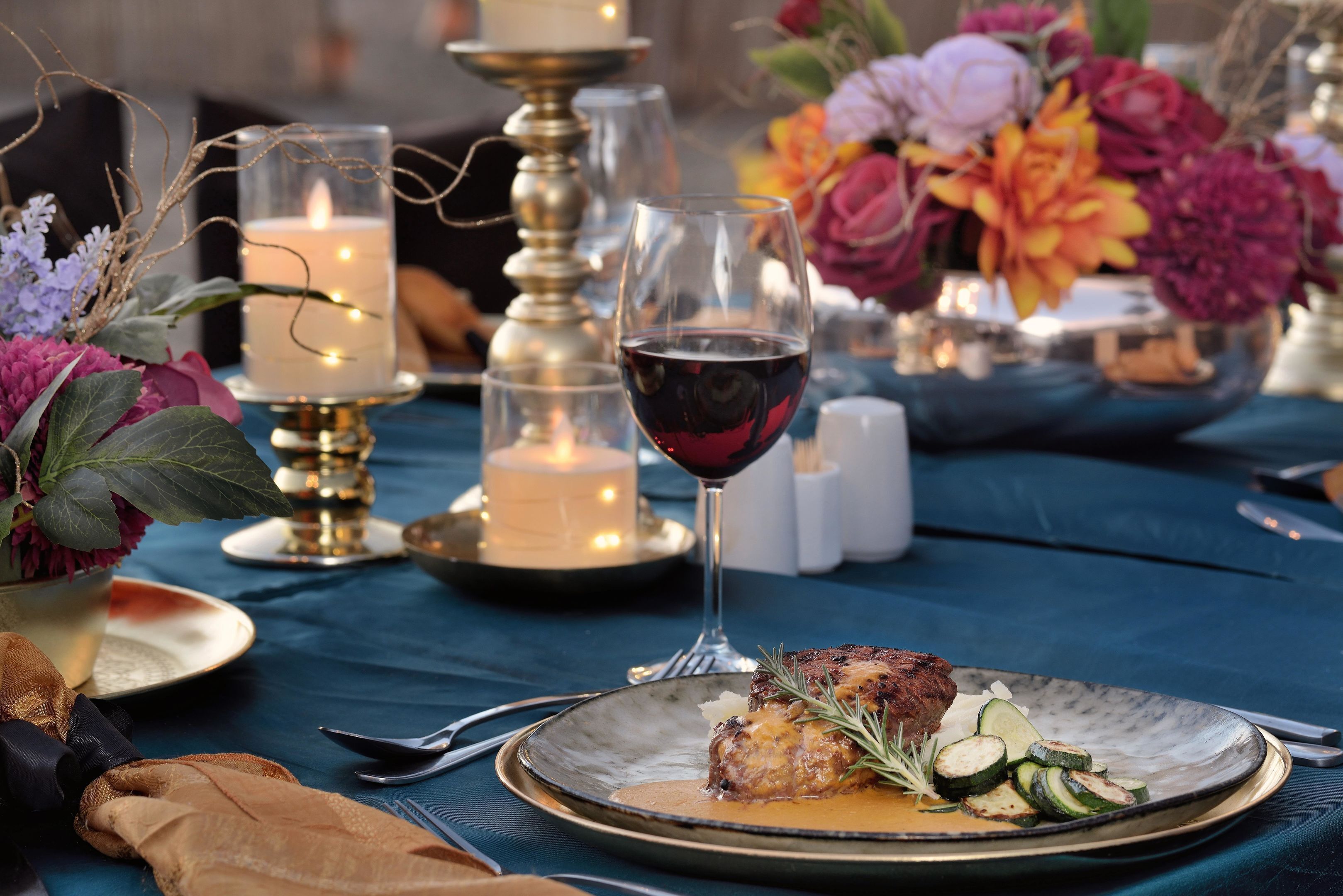 Una mesa dispuesta con un plato de comida, una copa de vino tinto, velas y un florero con flores.