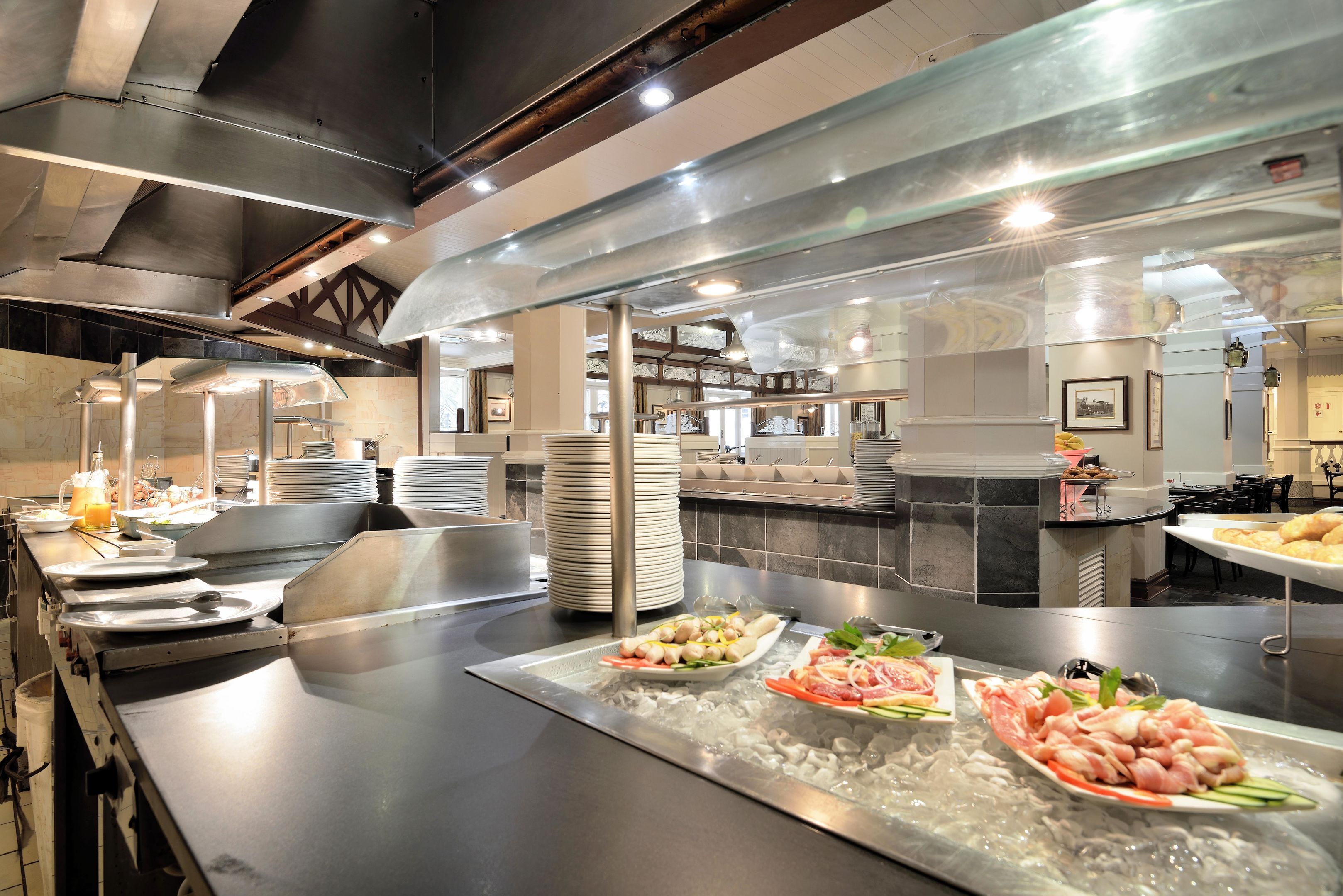 A buffet setup with various food items on a cold serving tray, surrounded by stacked plates and a stainless steel counter.