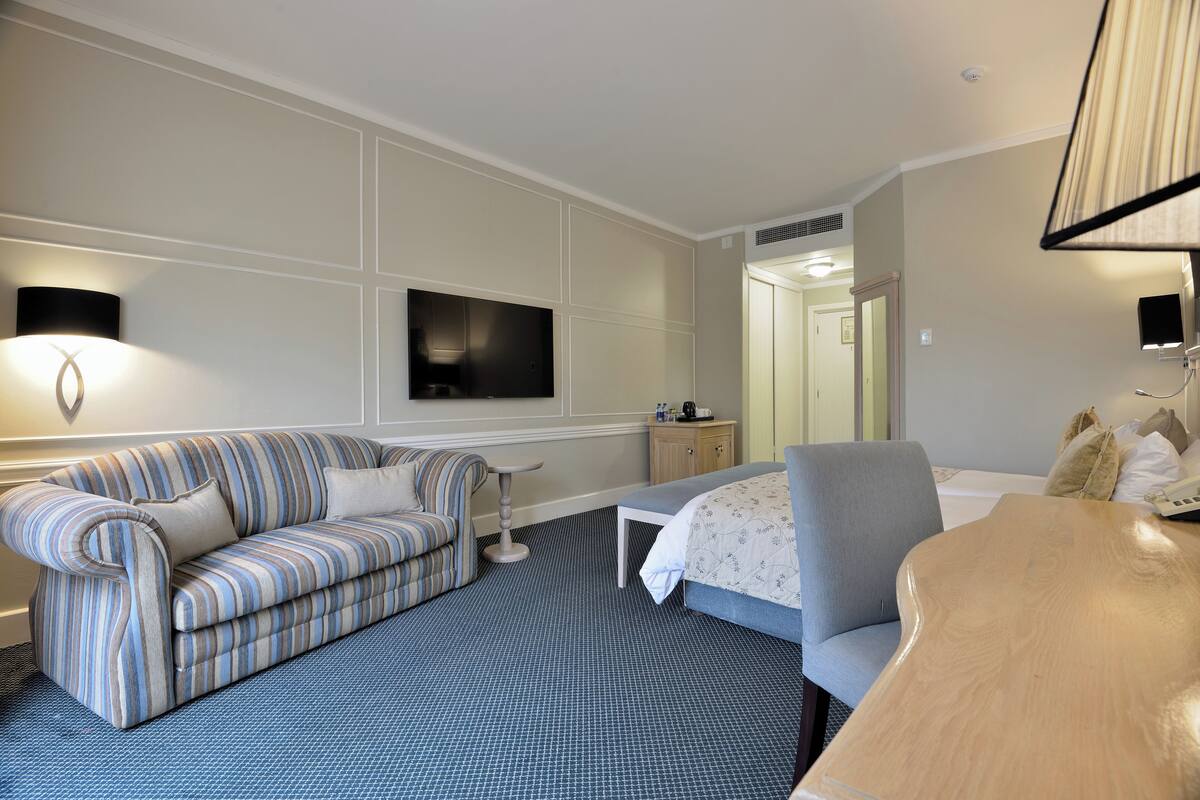 Une chambre d’hôtel avec un canapé à rayures, une petite table, un lit avec une tête de lit grise et un bureau en bois.