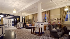 A spacious lobby with a patterned carpet, a reception desk, seating area with armchairs, and a large column. - Swakopmund Hotel (Swakopmund)