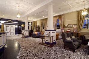 Een ruime lobby met een kamerbreed tapijt, een receptiebalie, een zithoek met fauteuils en een grote zuil.