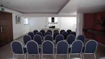Uma sala de conferências com fileiras de cadeiras voltadas para um púlpito, um letreiro com 'Contorno Hotel' na parede e uma escadaria à esquerda.