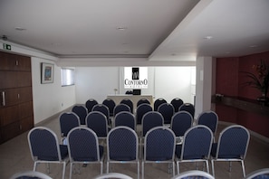 A conference room with rows of chairs facing a podium, a sign for 'Contorno Hotel' on the wall, and a staircase to the left.