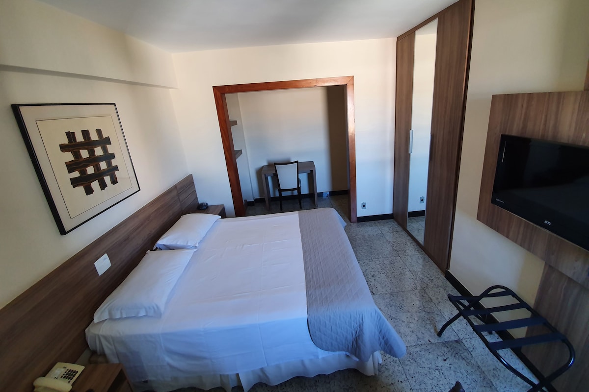 A hotel room with a bed, a desk, a chair, a TV, and a framed picture on the wall.