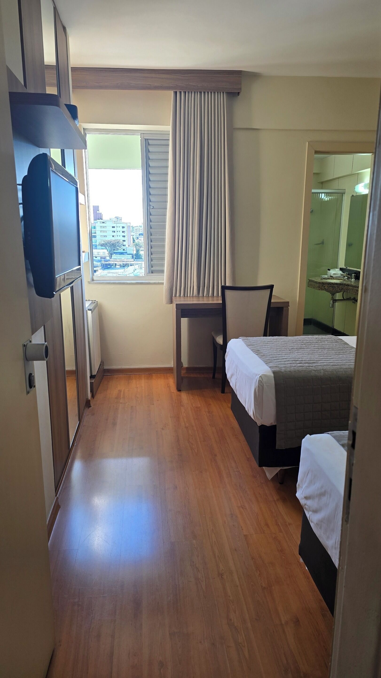 A hotel room with a wooden floor, a bed, a desk, a chair, a television, and a window with curtains.