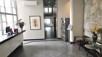 Uma área de recepção com balcão, obras de arte emolduradas, frigorífico e espaço de estar com um vaso de flores.