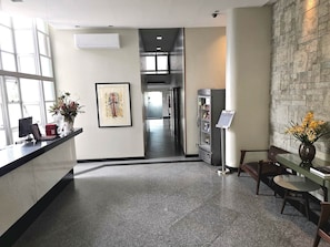 A reception area with a counter, framed artwork, a refrigerator, and a seating area with a vase of flowers.