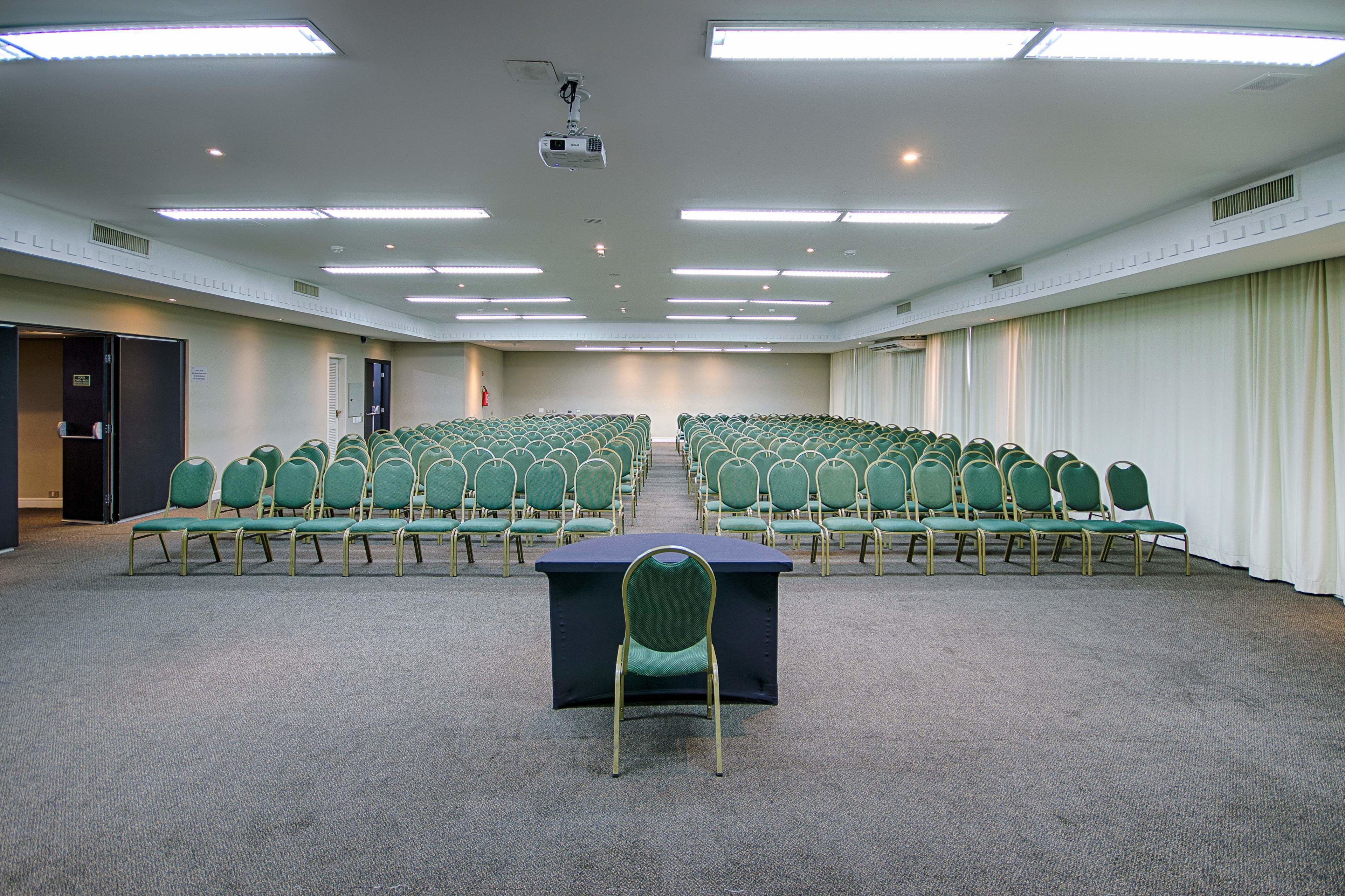 Een vergaderruimte met rijen groene stoelen, een centrale tafel en een projector aan het plafond.