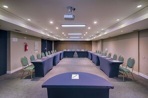 Een vergaderruimte met een lange tafel, groene stoelen en een beamer aan het plafond.