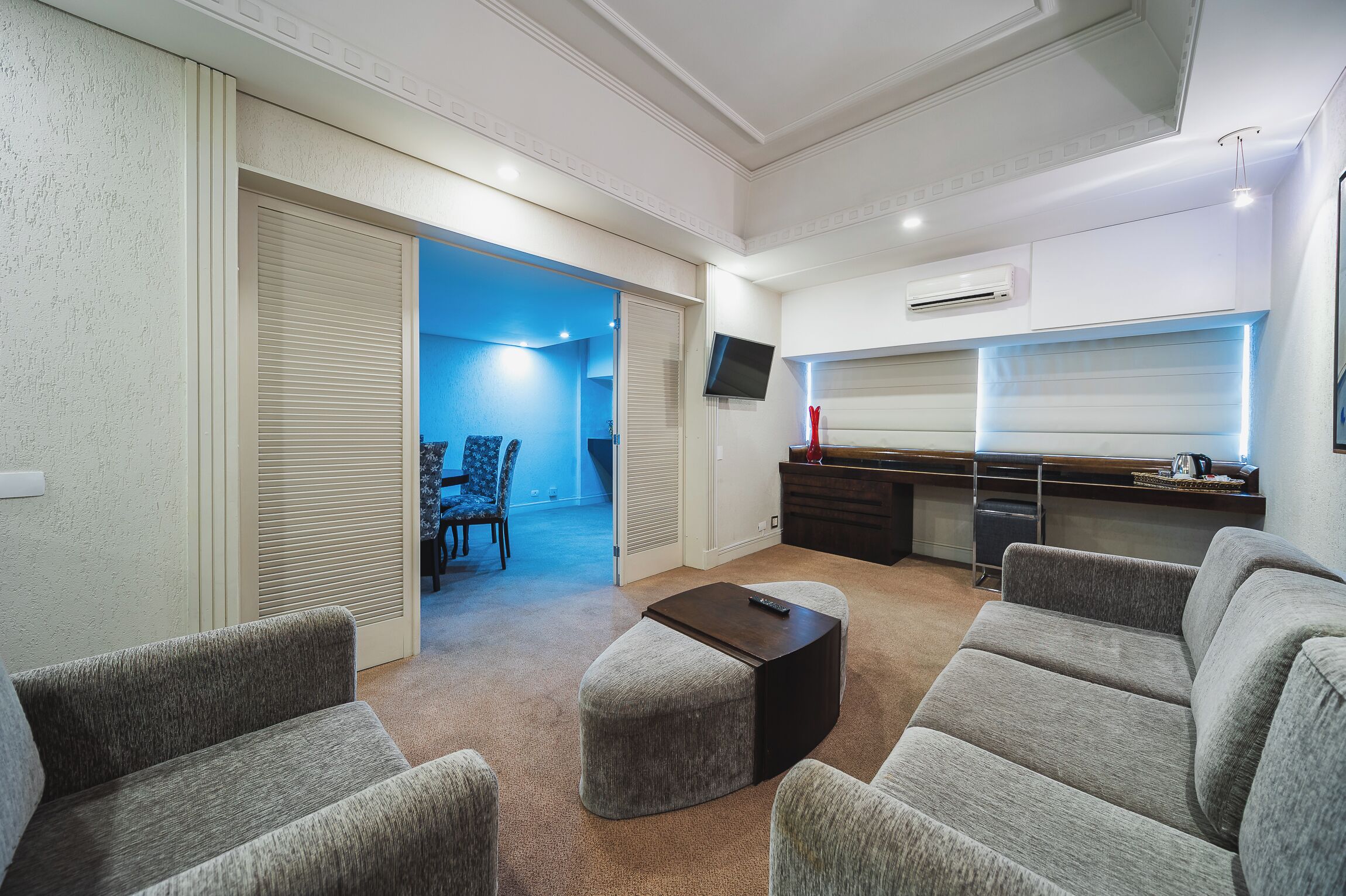 A modern living room with grey upholstered sofas, a dark coffee table, and a kitchen area with a TV and air conditioning unit.
