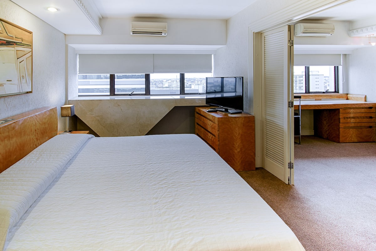 A hotel room with a large bed, a desk with a computer, a television, and a view of the city through the window.